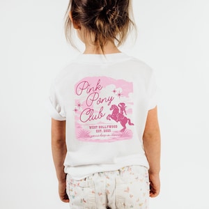 May include: A white t-shirt with a pink graphic on the back. The graphic features the text "Pink Pony Club" with a pink illustration of a person riding a horse. Additional text reads "West Hollywood Est. 2023" and "I'm gonna keep on dancing."