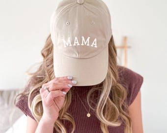 May include: A beige baseball cap with the word "MAMA" embroidered in white block letters across the front. The cap has a curved brim and a button on top. The cap is worn by a person with long, wavy blonde hair.