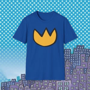 May include: A royal blue t-shirt featuring a gold crown design with a black outline. The crown is centered on the shirt. The background has a cityscape illustration and a blue and white polka dot pattern.