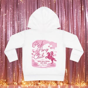 May include: White hoodie with a pink graphic on the back. The graphic reads "Pink Pony Club" with a silhouette of a person riding a horse. The text "West Hollywood Est. 2023" is also included. The hoodie has a hood and long sleeves.