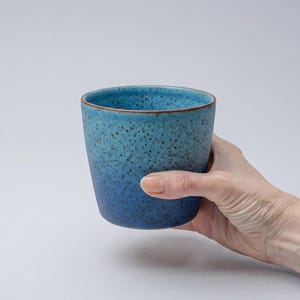 Può includere: Una tazza di ceramica blu con una texture maculata e un effetto sfumato, che passa da un blu più scuro alla base a un blu più chiaro sul bordo. La tazza ha un bordo marrone ed è tenuta da una mano.