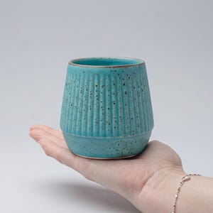 Op de afbeelding: Een turquoise keramische beker met verticale geribbelde details. De beker heeft een afgeronde basis en een licht uitlopende rand. Het oppervlak heeft een gespikkelde textuur. De beker wordt in een hand gehouden tegen een effen achtergrond.