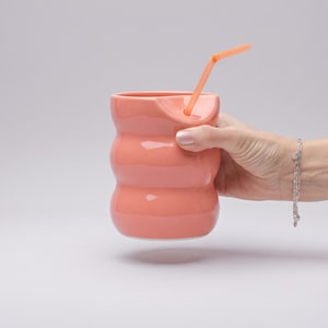 Può includere: Un bicchiere in ceramica color corallo con una superficie esterna ondulata e una finitura liscia. Il contenitore presenta una tacca distinta lungo il bordo superiore e include una cannuccia di plastica abbinata inserita nell'apertura.
