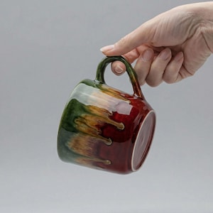 Könnte beinhalten: Eine Keramiktasse mit abgerundeter Form und Henkel. Die Tasse hat eine Glasur mit einer Mischung aus Grün, Braun und Rot. Die Tasse wird von einer Hand vor einem schlichten Hintergrund gehalten.