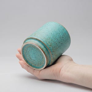 Puede incluir: Un vaso de cerámica turquesa con una textura moteada. La taza cilíndrica tiene una base ligeramente ensanchada y un borde marrón contrastante. La cerámica hecha a mano se sostiene en una mano, mostrando su tamaño y forma. La taza es adecuada para bebidas.