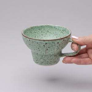 Pode incluir: Uma caneca de cerâmica larga e rasa com um esmalte verde-menta salpicado de manchas castanho-escuras. A caneca apresenta uma silhueta escalonada distinta, uma borda lisa e uma pequena alça curva que se estende na lateral do recipiente.