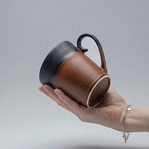 Könnte beinhalten: Eine zweifarbige Keramik-Tasse mit schwarzem Oberteil und braunem Korpus. Sie hat einen gebogenen Henkel und eine weiße Innenseite. Die Tasse wird in einer Hand vor grauem Hintergrund gehalten.