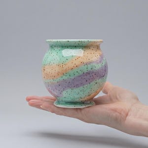 Puede incluir: Un pequeño jarrón de cerámica redondo con base de pedestal. El jarrón presenta un diseño en espiral en colores pastel: verde menta, melocotón y lavanda, salpicado de puntos negros. El jarrón se sostiene en una mano sobre un fondo gris.