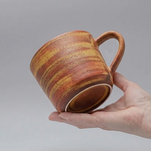 以下が含まれることがあります： テクスチャ加工された表面と茶色と黄色のストライプ模様のセラミックマグ。 マグには湾曲したハンドルがあり、手で持っています。 マグの高さは約10cmです。