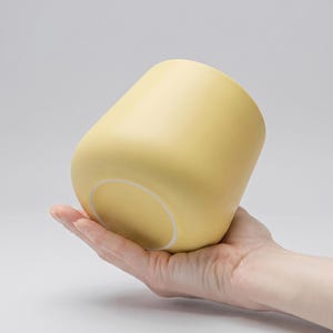 Puede incluir: Una taza de cerámica amarillo pálido con forma redondeada y cónica. La taza se sostiene en una mano, mostrando su acabado liso y mate y su interior blanco. El diseño de la taza es simple y moderno, adecuado para bebidas.