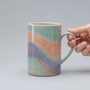 Op de afbeelding: Een hoge keramische mok met een gebogen handvat. De mok heeft een gespikkelde, lichtgroene basiskleur met diagonale strepen in oranje, paars en lichtgroen. De mok wordt door een hand vastgehouden.