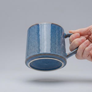 Può includere: Una tazza di ceramica blu con manico. La tazza ha una superficie strutturata e un bordo marrone. La tazza è tenuta da una mano su uno sfondo grigio.