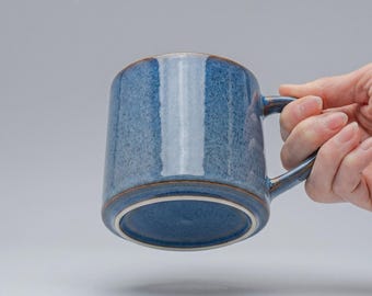 May include: A blue ceramic mug with a handle. The mug has a textured surface and a brown rim. The mug is held by a hand against a gray background.