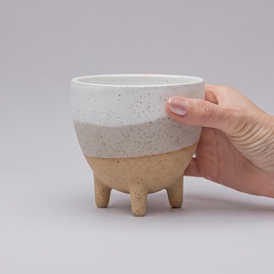 Puede incluir: Una maceta de cerámica con tres patas cortas y textura moteada. La parte superior es blanca, pasando por una sección media de color beige claro hasta una base de color tostado. El recipiente tiene una forma redondeada y un borde liso.