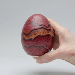 Può includere: Un oggetto decorativo a forma di uovo con una superficie strutturata. L'uovo presenta fasce di tonalità rosse, marroni e arancioni. L'oggetto è tenuto in mano su uno sfondo neutro. L'uovo è alto circa 10 cm.