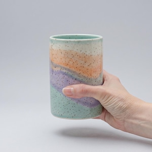 Könnte beinhalten: Ein zylindrischer Keramikbecher mit einem gesprenkelten, geschichteten Design. Die Farben umfassen Mintgrün, Lavendel, Orange und Off-White. Der Becher wird von einer Hand gehalten, die seine glatte, bemalte Oberfläche zeigt. Der Becher ist etwa 13 cm hoch.