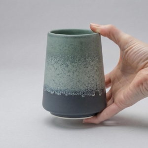 Può includere: Un bicchiere in ceramica dalla forma cilindrica affusolata con una finitura opaca liscia. L'esterno presenta una sfumatura di colore che passa da un grigio antracite scuro alla base a un verde salvia chiaro testurizzato sul bordo superiore.
