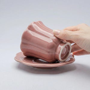 Könnte beinhalten: Ein staubrosa Teetassen- und Untertassen-Set. Die Teetasse hat ein einzigartiges, geriffeltes Design und liegt verkehrt herum auf der passenden Untertasse. Das Set ist aus Keramik und hat eine glänzende Oberfläche. Der Hintergrund ist einfarbig und neutral.