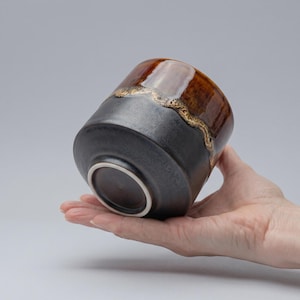 Puede incluir: Taza de cerámica hecha a mano con base gris oscuro y parte superior marrón brillante. El borde presenta un adorno decorativo de color dorado. La taza se sostiene en una mano, mostrando su forma y diseño.