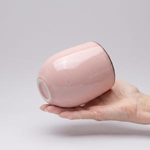 Può includere: Un bicchiere in ceramica rosa chiaro liscio con una silhouette arrotondata senza manico. Il recipiente presenta una finitura lucida e un bordo scuro a contrasto. La base non è smaltata, rivelando il materiale ceramico bianco sotto lo smalto.