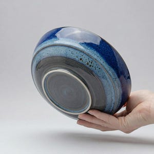 Pode incluir: Uma tigela de cerâmica com uma cobertura azul brilhante e em camadas. O exterior da tigela apresenta faixas de azul escuro e claro, com um interior azul escuro. A tigela é segurada em uma mão, mostrando sua forma e design.