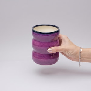 Könnte beinhalten: Ein zylindrischer Keramikbecher mit einer leuchtend violetten Glasur und einem dunkelblauen Rand. Der Becher weist eine ausgeprägte, dreistufige, wellenförmige oder blasige Außenstruktur auf. Das Innere des Bechers ist glatt und cremefarben.