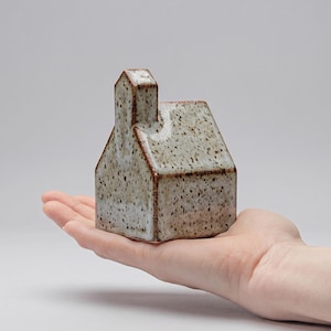 Può includere: Piccola statuetta a forma di casa in ceramica con camino. La casa è macchiata di macchie marroni e ha un colore beige chiaro. I bordi sono marrone scuro. La statuetta è tenuta in una mano.