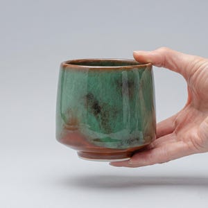 Może przedstawiać: Ceramiczny kubek o gładkiej, zaokrąglonej sylwetce z błyszczącym szkliwem. Zewnętrzna strona prezentuje mieszankę żywych odcieni turkusu i zieleni, przechodzących u podstawy w ciepły, ziemisty brąz, z subtelnymi ciemnymi nakrapianymi akcentami.