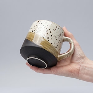 Puede incluir: Una taza de cerámica con un diseño único. La taza presenta una base negra, una banda diagonal verde oliva y una sección superior blanca moteada. El asa también es blanca moteada. La taza se sostiene en ángulo, mostrando su diseño.