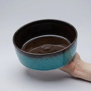 以下が含まれることがあります： 濃い茶色の内側とターコイズブルーの外側を持つセラミックボウル。ボウルは手で持たれており、その丸い形と艶出し仕上げを示しています。ボウルの縁は、より濃い茶色です。