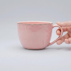 Può includere: Una tazza in ceramica rosa con un corpo arrotondato e un manico grande. La tazza ha una finitura liscia e lucida e un colore rosa chiaro. Il bordo della tazza ha un colore più scuro e contrastante. La tazza è tenuta da una mano.