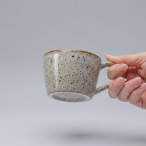 May include: A ceramic mug with a speckled light gray and beige finish, featuring a smooth, rounded body and a small handle. The rim of the mug has a thin, distinct brown edge, and the overall surface is covered in dark brown speckles.