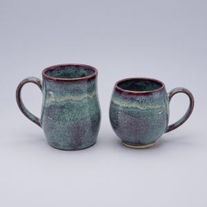 May include: Two ceramic mugs with a reactive glaze finish in shades of deep purple, teal, and cream. The taller mug features a cylindrical body with a curved handle, while the shorter mug has a rounded, bulbous shape and a smaller handle.