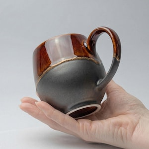Puede incluir: Una taza de cerámica hecha a mano con un esmalte de dos tonos. La mitad superior es de un marrón brillante, mientras que la mitad inferior es gris mate. Una fina banda dorada separa los dos colores. El asa es marrón y gris. La taza se sostiene en una mano.