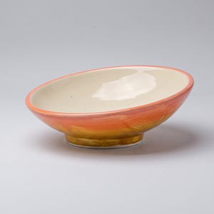 Può includere: Una ciotola in ceramica poco profonda con base arrotondata e bordo ampio e aperto. L'interno è di un colore bianco sporco liscio, mentre l'esterno presenta una vivace sfumatura dipinta a mano che passa dall'arancione caldo in alto al marrone scuro alla base.