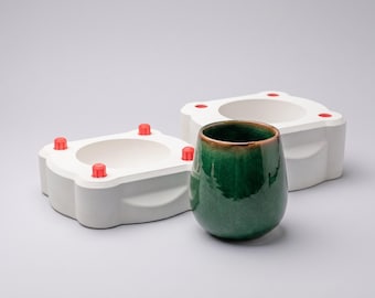 May include: A glossy, dark green ceramic cup with a subtle crackle glaze finish stands in front of two white plaster casting molds. Each mold features a square-shaped base with rounded corners and four small, bright red plastic alignment pins.