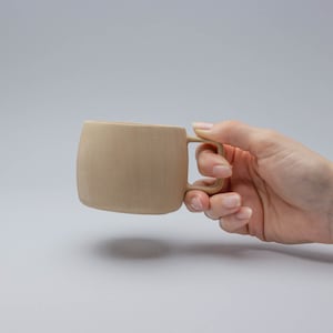 Könnte beinhalten: Eine beige Keramiktasse mit abgerundeter Form und einem einfachen Henkel. Die Tasse wird von einer Hand gehalten.