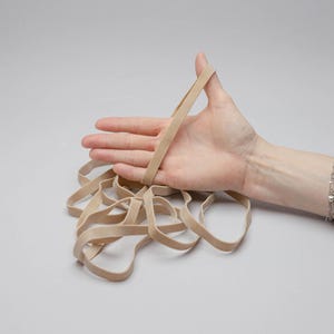 Puede incluir: Una mano sosteniendo una goma elástica beige, con varias otras gomas elásticas esparcidas debajo. Las gomas elásticas son de un color neutro, y el fondo es gris claro liso.