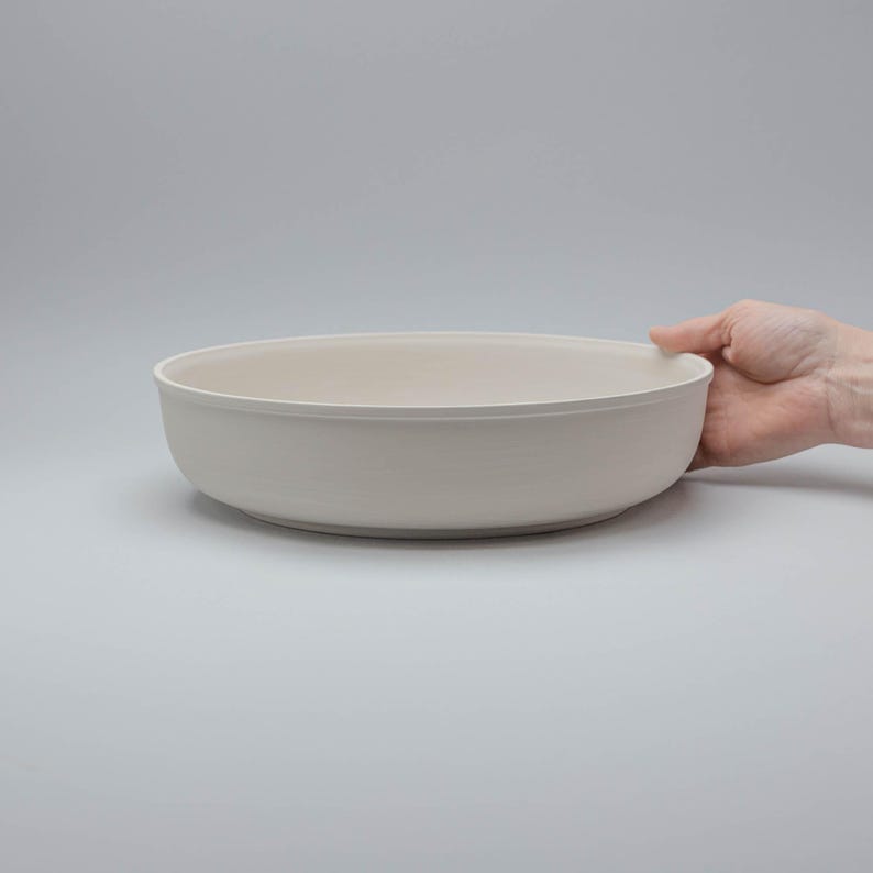 Pode incluir: Uma tigela grande de cer&acirc;mica branca suja com um design largo e raso. A tigela &eacute; segurada por uma m&atilde;o em um fundo cinza claro liso. A borda da tigela &eacute; ligeiramente curvada e o interior &eacute; um tom mais claro de branco sujo.