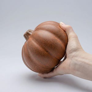 May include: A brown ceramic pumpkin with a textured surface and a stem. The pumpkin is held in a hand, showcasing its size and shape. This decorative item is perfect for fall and autumn-themed home decor.