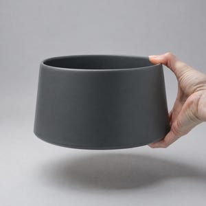 Könnte beinhalten: Eine dunkelgraue Keramikschale mit einer breiten, leicht ausgestellten Form. Die Schale hat eine glatte, matte Oberfläche und wird von einer Hand gehalten. Das Design der Schale ist schlicht und modern, geeignet zum Servieren oder Ausstellen.