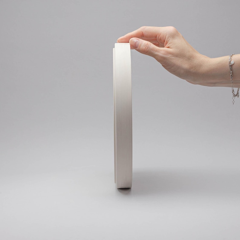 May include: A circular, matte white object with a smooth, flat edge and a slightly recessed centre. The object is positioned vertically, showcasing its thin profile and uniform, clean finish. The design is simple, featuring a solid, neutral colour.