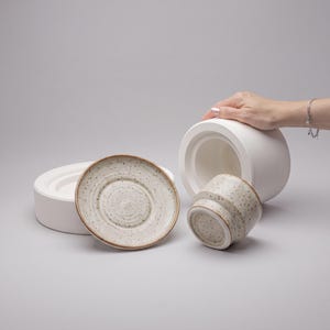 Peut inclure: Un ensemble d'ustensiles de cuisine en céramique comprenant un grand récipient cylindrique blanc mat, une assiette circulaire mouchetée avec un bord brun et une petite tasse mouchetée à base conique, disposés sur un fond clair uni.