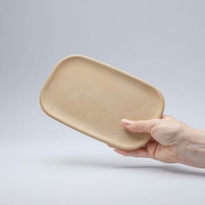 以下が含まれることがあります： 丸みを帯びた縁のある薄い茶色の木製トレイ。トレイは手で持たれています。