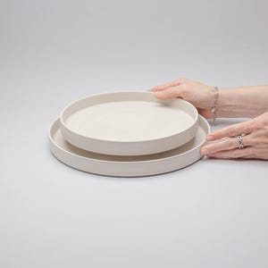 May include: Two stacked circular plates with a matte off-white finish. The plates feature a flat base and raised vertical edges, creating a simple, uniform profile. The surface of the ceramic material appears smooth and consistent in colour.