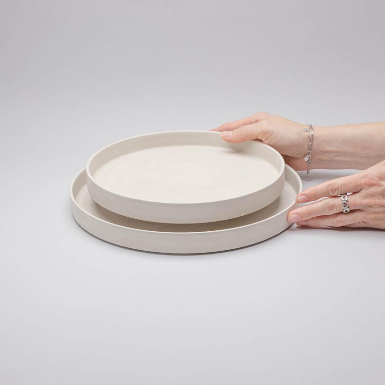 May include: Two stacked circular plates with a matte off-white finish. The plates feature a flat base and raised vertical edges, creating a simple, uniform profile. The surface of the ceramic material appears smooth and consistent in colour.