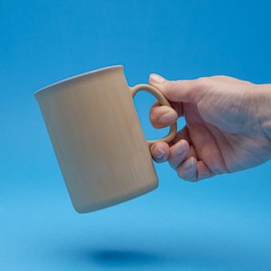 Könnte beinhalten: Eine beige Keramiktasse mit einem hellvioletten Rand und einem Henkel. Die Tasse wird von einer Hand vor einem blauen Hintergrund gehalten.