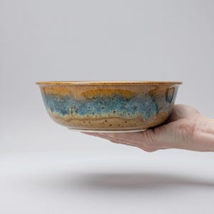 Puede incluir: Un cuenco de cerámica hecho a mano con un esmalte único. El cuenco presenta una mezcla de colores marrón, azul y beige. El cuenco es sostenido por una mano, mostrando su tamaño y forma. Ideal para servir alimentos o como objeto decorativo.