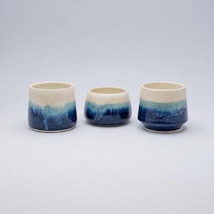 Op de afbeelding: Drie keramische bekers met een glad, crèmekleurig bovendeel en een diepblauwe, druipende glazuurafwerking aan de onderkant. De vaten hebben een afgeronde, taps toelopende vorm met een brede opening en een platte bodem met een kleurverloop.