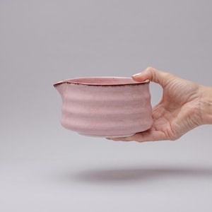 Può includere: Una ciotola in ceramica rosa chiaro con una texture a coste orizzontali e un piccolo beccuccio. Il bordo della ciotola presenta una sottile linea di accento marrone scuro, in contrasto con il morbido smalto rosa pastello esterno.